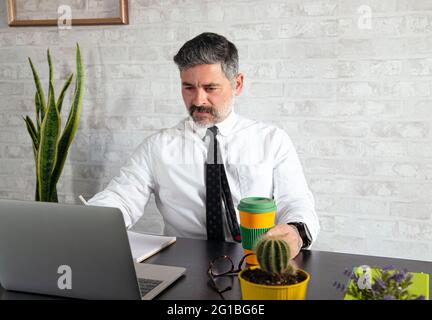 Middle aged male entrepreneur in formal wear working on netbook at table with hot drink to go Stock Photo
