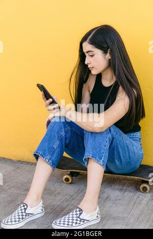 Young female skater surfing internet on cellphone while sitting on skateboard on walkway in daytime Stock Photo