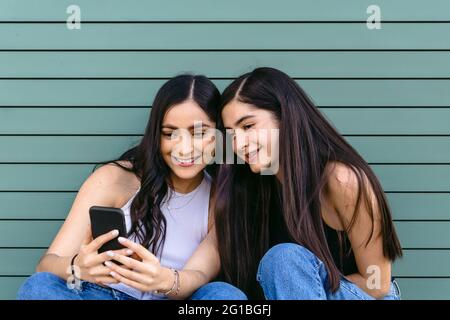 Content female siblings with long hair surfing internet on mobile phone while spending time together in daylight Stock Photo