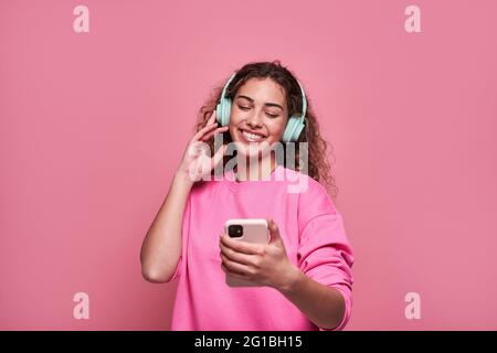 Cheerful teenager with curly hair scrolling mobile phone while listening to music standing against pink background Stock Photo