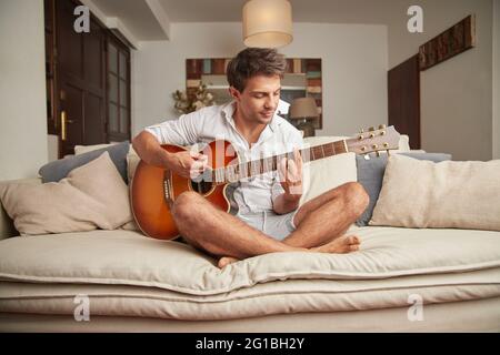 Adult male in casual clothes sitting on couch playing acoustic guitar in light living room Stock Photo