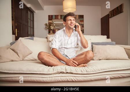 Adult male in casual clothes sitting on couch with pillows while having conversations on smartphone in light living room while looking away Stock Photo