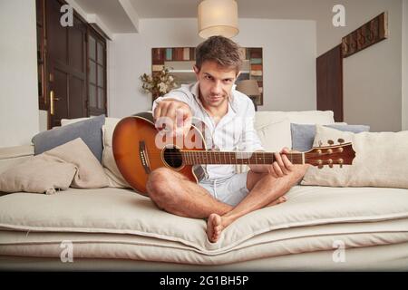 Adult male in casual clothes sitting on couch with acoustic guitar in hands while pointing finger at camera in light living room and looking at camera Stock Photo