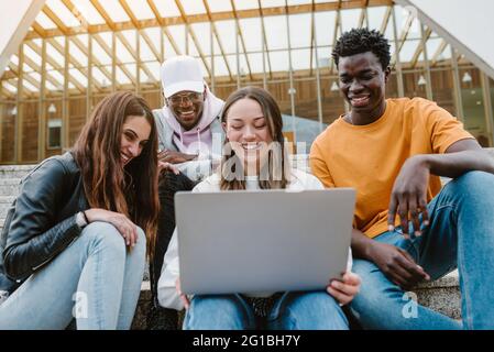 Diverse young friends smiling widely while watching video on netbook together while sitting on concrete stairs Stock Photo