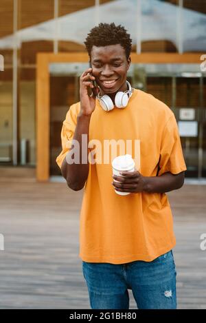 Smiling African American male with cup of coffee laughing happily while speaking on mobile phone and looking away Stock Photo