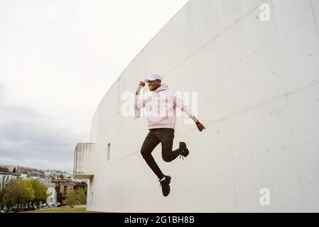 Low angle of full body of energetic African American male in casual wear laughing happily with fist up celebrating victory while jumping Stock Photo