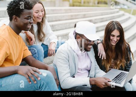 Diverse young friends smiling widely while watching video on netbook together while sitting on concrete stairs Stock Photo