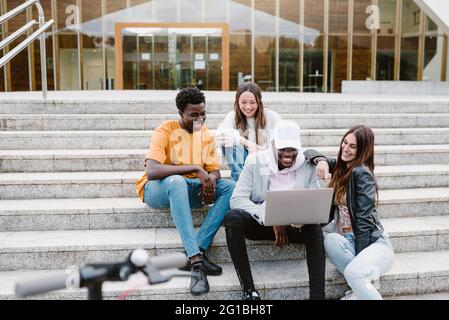 Diverse young friends smiling widely while watching video on netbook together while sitting on concrete stairs Stock Photo