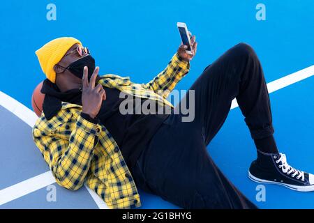 From above stylish African American male in face mask lying on basketball court and taking selfie on smartphone while showing V sign Stock Photo