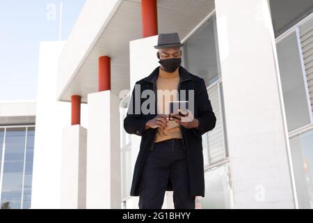Low angle positive African American male in stylish wear and face mask browsing tablet while standing on sunny street Stock Photo