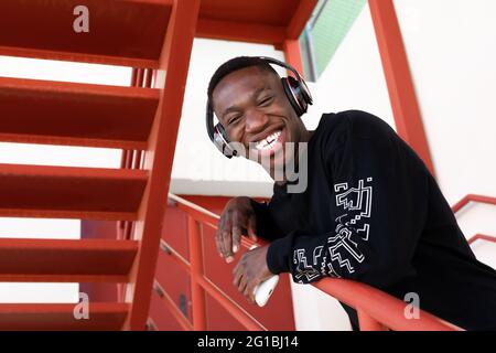 Cheerful African American male in casual wear and wireless headphones smiling looking at camera while standing on metal staircase Stock Photo