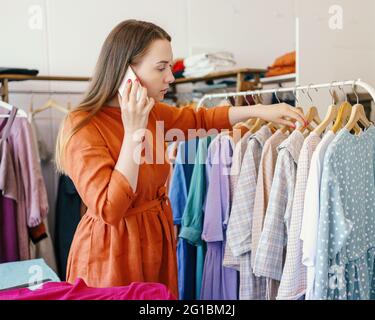 Young woman owner of fashion clothing store or studio standing near designer dresses hanging on clothes rack and taking order from client on mobile phone. Female dressmaker working in workshop Stock Photo