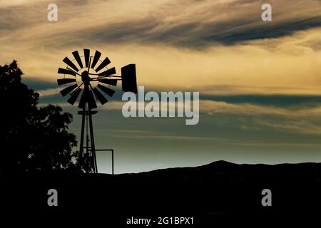 Rich, dark silhouette of a vintage windmill against yellow clouds and a bluish sky just as twilight takes hold in America's old west. Stock Photo