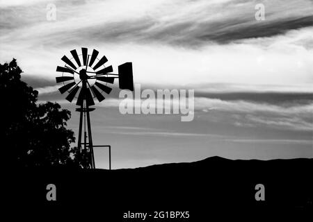 Rich, dark, black and white silhouette of a vintage windmill against an overcast, clouded sky just as twilight takes hold in America's old west. Stock Photo