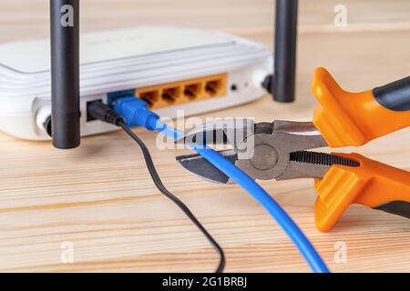 Wire snippers cutting the blue network cable connected to white Wi-Fi wireless router on a wooden desk. Prohibit and restrict access to the internet. Stock Photo