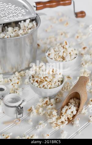 Popcorn pan maker, white bowls and wooden scoop full of popcorn. Messy food scene, in white. Retro, vintage ambience. Stock Photo