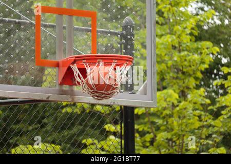 close up of a basketball going through the net of the basketball hoop with glass backboard visible and trees in background, a score in basketball game Stock Photo