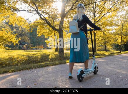 Young blonde woman with backpack is going to ride an electric scooter from the park in early autumn Stock Photo