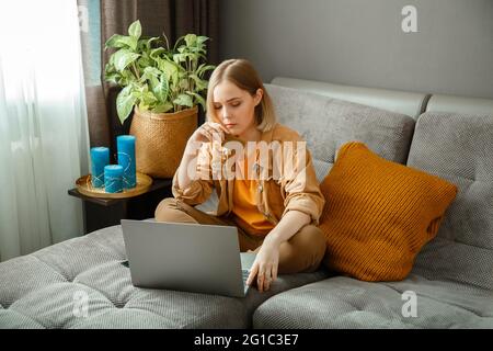 Beautiful young woman shopper makes online purchases using laptop and credit debit cards while sitting on couch at home. Woman make online purchases Stock Photo