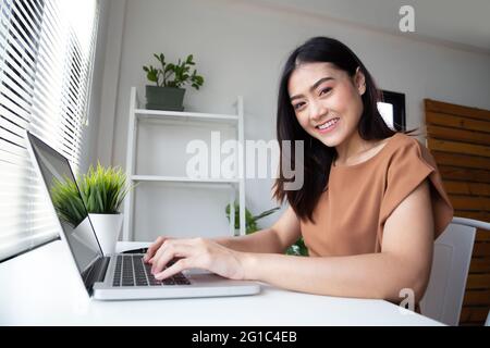 Young Asian woman in casual clothing using laptop computer checking email and smiling while working indoors at living room. Working from home or stay Stock Photo