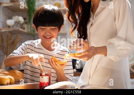 Smiling mother and son cooking and making breakfast food with bread and jam strawberry together in the kitchen at home. Happy life activity concept Stock Photo