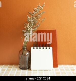 Notebooks and vase of misty blue dry flowers on beige ceramic mosaic tile desk. Orange background. Work and study place Stock Photo