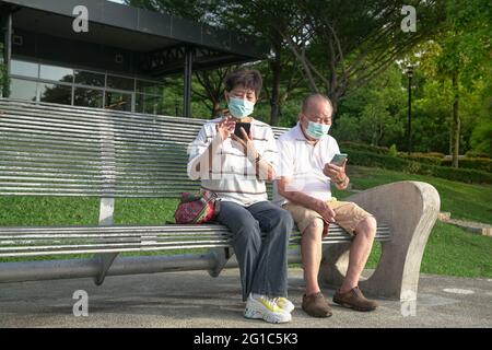 Senior Asian couple wearing face mask, sitting and reading their phones in a park. Digital lifestyle during pandemic. Stock Photo