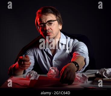 Unshaved man in shirt, jacket and glasses at work, sits behind desk, table with books and sheets and crumpled paper Stock Photo