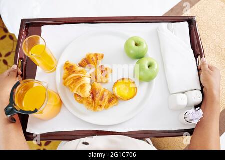 Maid carrying tray with breakfast to hotel room, it consist of fresh croissants, apples, and glasses of juice, view from above Stock Photo
