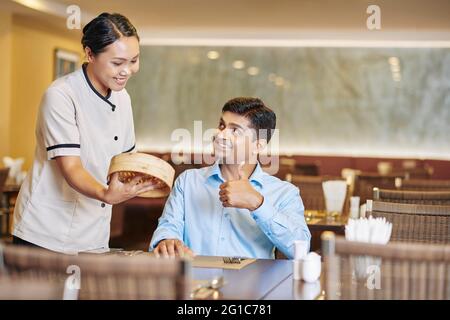 Smiling waitress giving serving plate with delicious baozi, Chinese steamed meat buns to customer sitting at table and showing thumbs-up Stock Photo