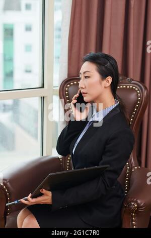 Young serious businesswoman sitting in armchair with documents folder and talking on phone Stock Photo