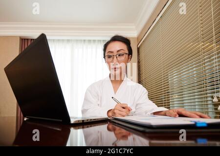 Serious young female entrepreneur reading document on laptop screen and analyzing sales report Stock Photo