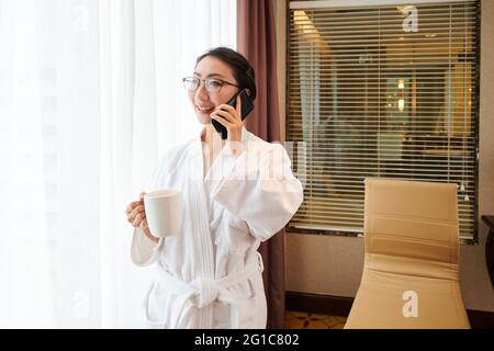 Happy young woman in bathrobe drinking morning coffee and talking on phone Stock Photo