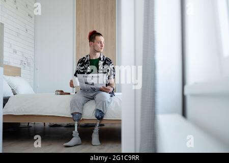 Portrait of disabled young man reading newspapers on bed indoors at home, leg prosthetic concept. Stock Photo