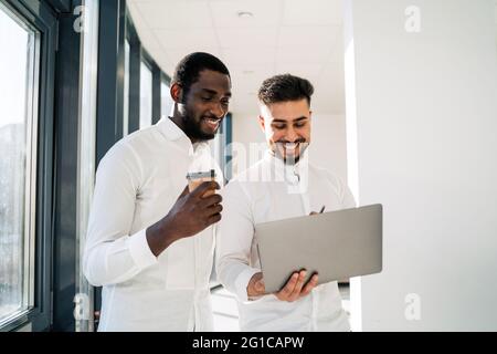Two multiracial cheerful corporate employees are talking while standing with laptop in the middle of the office Stock Photo