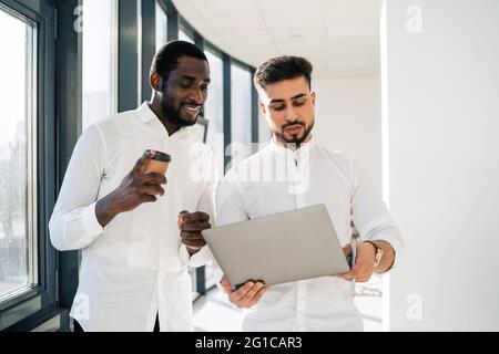 Two interracial businessmen talking while standing in the office with laptop Stock Photo