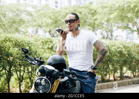 Man in sunglasses standing next to his motorcycle and recording voice message for friend or coworker Stock Photo