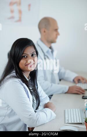 Smiling young doctor sitting at desk next to his colleague and working on laptop, filling patients data Stock Photo