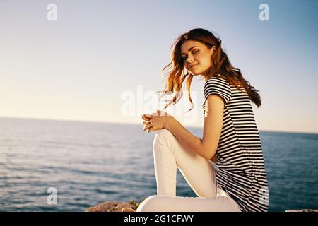 a woman in a striped T-shirt and white trousers sits on a stone near the sea in summer Stock Photo