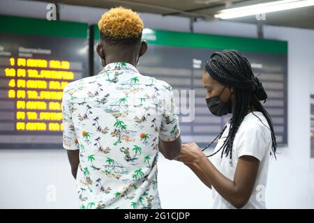 Back view of couple in airport wearing protective face masks, looking at their smartphone and the travel board with flights information. Stock Photo