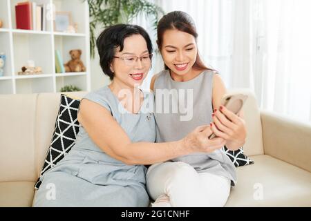 Elegant smiling senior woman and her adult daughter sitting on sofa at home and taking selfie Stock Photo