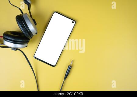 Modern smartphone with white mockup on screen with closed headphones with jack on yellow background with copy space, top view. Template for music apps Stock Photo
