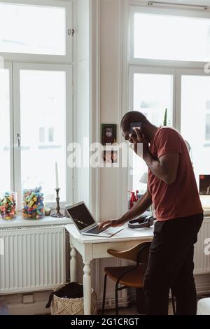 Side view of businessman talking on smart phone while using laptop at home Stock Photo