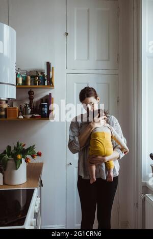 Mother carrying baby boy while standing in kitchen Stock Photo
