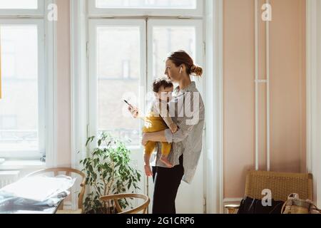 Businesswoman carrying baby boy while holding smart phone at home Stock Photo