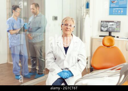 Portrait of dentist senior woman waiting for sick patient in stomatology hospital office during medical examination. Stomatology nurse writing teethcare treatment on clipboard inviting man in cabinet Stock Photo