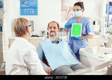 Nurse dentist showing green screen display to stomatology senior doctor while examining tooth pain to man patient sittinh on dental chair. Woman examinating using mock up chroma key tablet Stock Photo