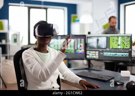African video editor experiencing VR goggles, gesturing, editing video film montage working with footage and sound on computer with two displays. Videographer processing movie in creative agency Stock Photo