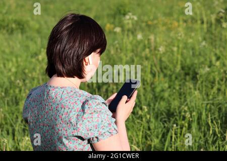 Woman in wireless headset using smartphone on a summer meadow. Female hands with mobile phone on green nature background Stock Photo