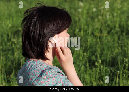 Woman touches wireless earphone in the ear standing on summer meadow. Headset, listening to music and voice call on a nature Stock Photo